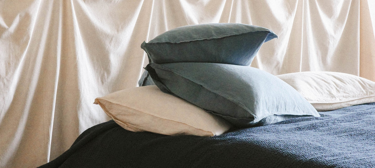 A stack of two blue pillows resting on a bed with a dark blue textured blanket, set against a backdrop of draped off-white fabric curtains.