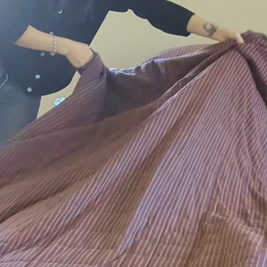 A person holding up a large quilt with thin vertical stripes in a muted reddish-brown and light beige color, set against a plain indoor wall.