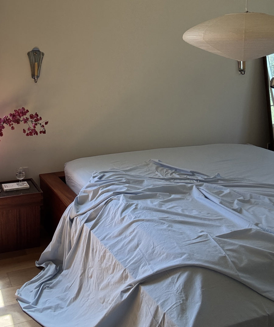 A bed with light blue and white bedding in a softly lit bedroom, with a wooden bedside table, wall sconce, and hanging pendant light visible.
