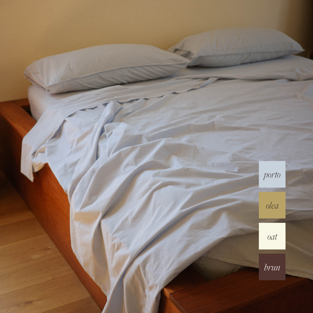 Light blue bed sheets and pillows on an unmade wooden bed frame in a bedroom setting, with a vertical color swatch overlay on the right side showing color options labeled porto, olea, oat, and brun.