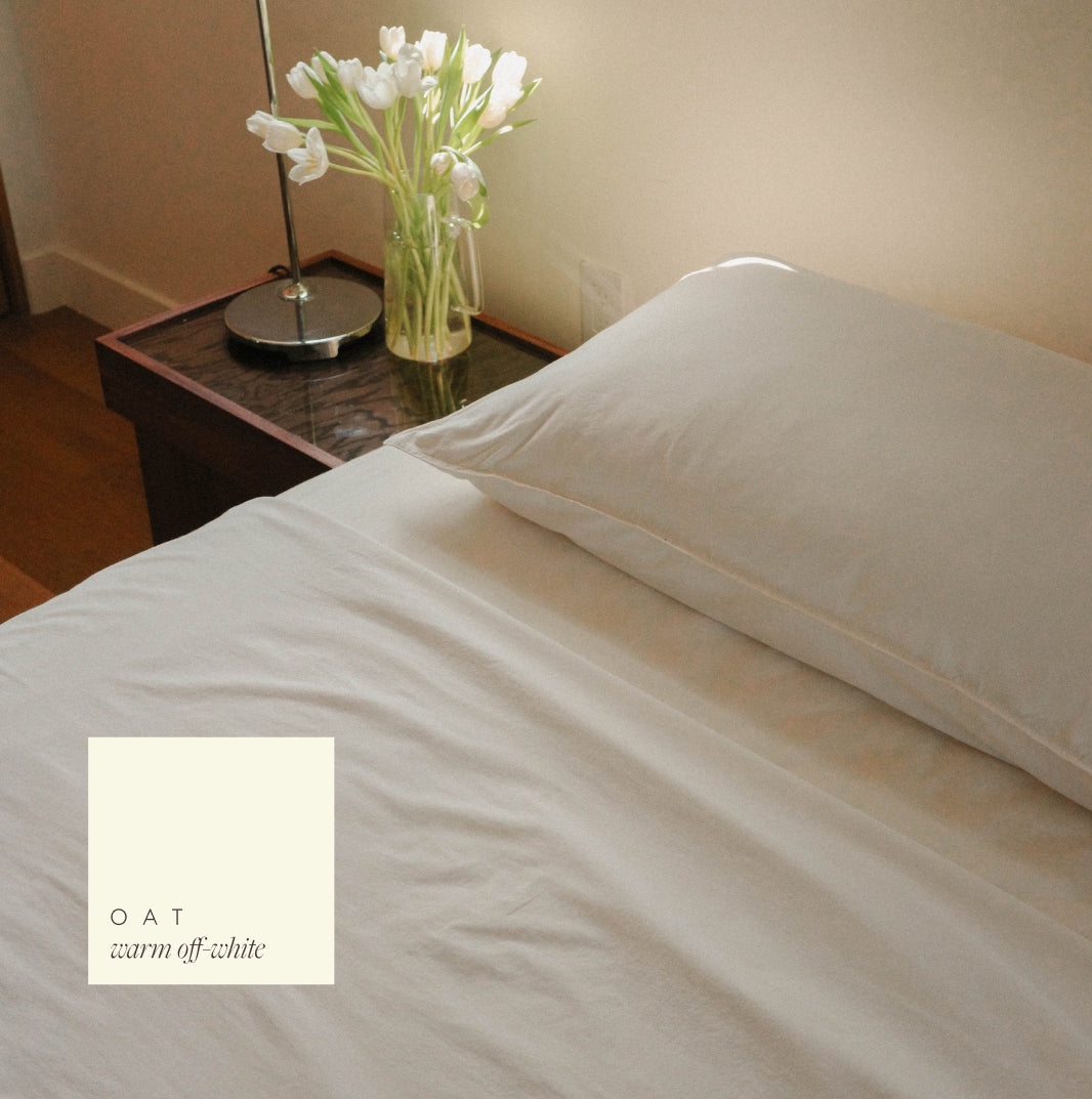 A close-up of a bed dressed in warm off-white bedding, with a matching pillow and a small cream color swatch labeled 'OAT warm off-white' in the lower left.