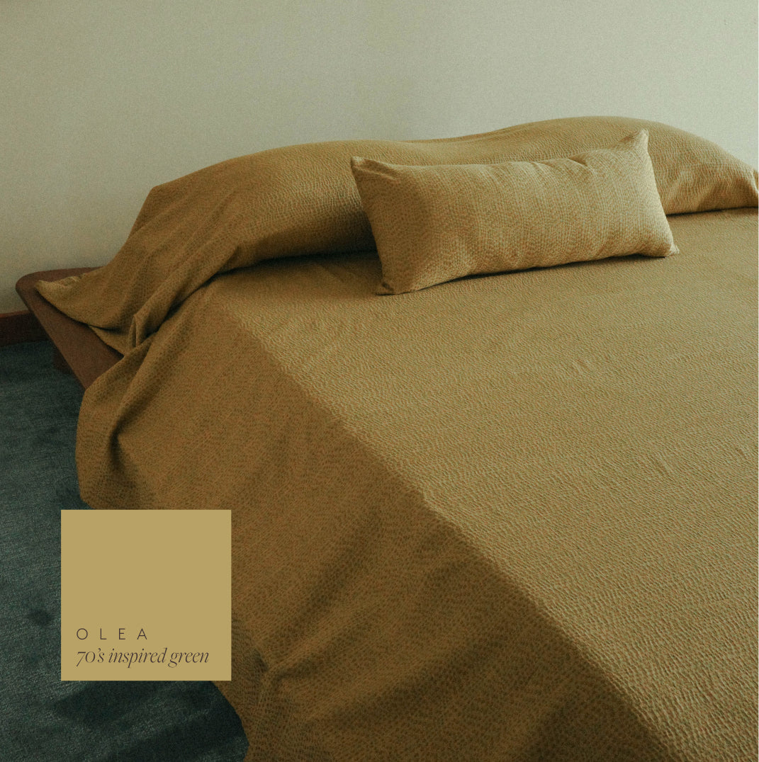 A bed dressed in textured olive-gold bedding with matching pillowcases, shown in a softly lit room. A square color label in the lower left reads 'OLEA 70's inspired green'.