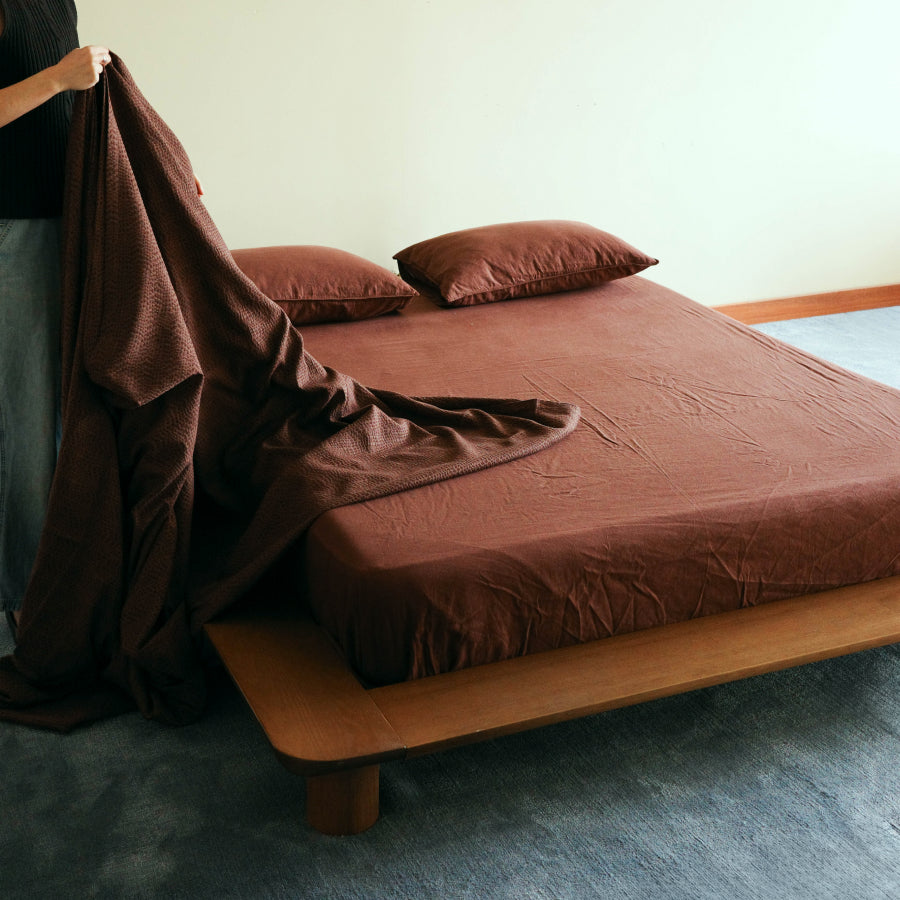 A person is pulling a dark brown textured sheet off a bed with matching brown pillows and sheets, set in a minimalist bedroom with a wooden platform bed frame.