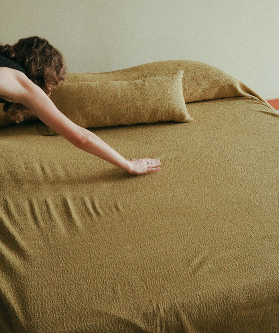 A person reaches across a bed to smooth out a textured olive-brown bedding set, which includes a duvet cover and matching pillowcases.
