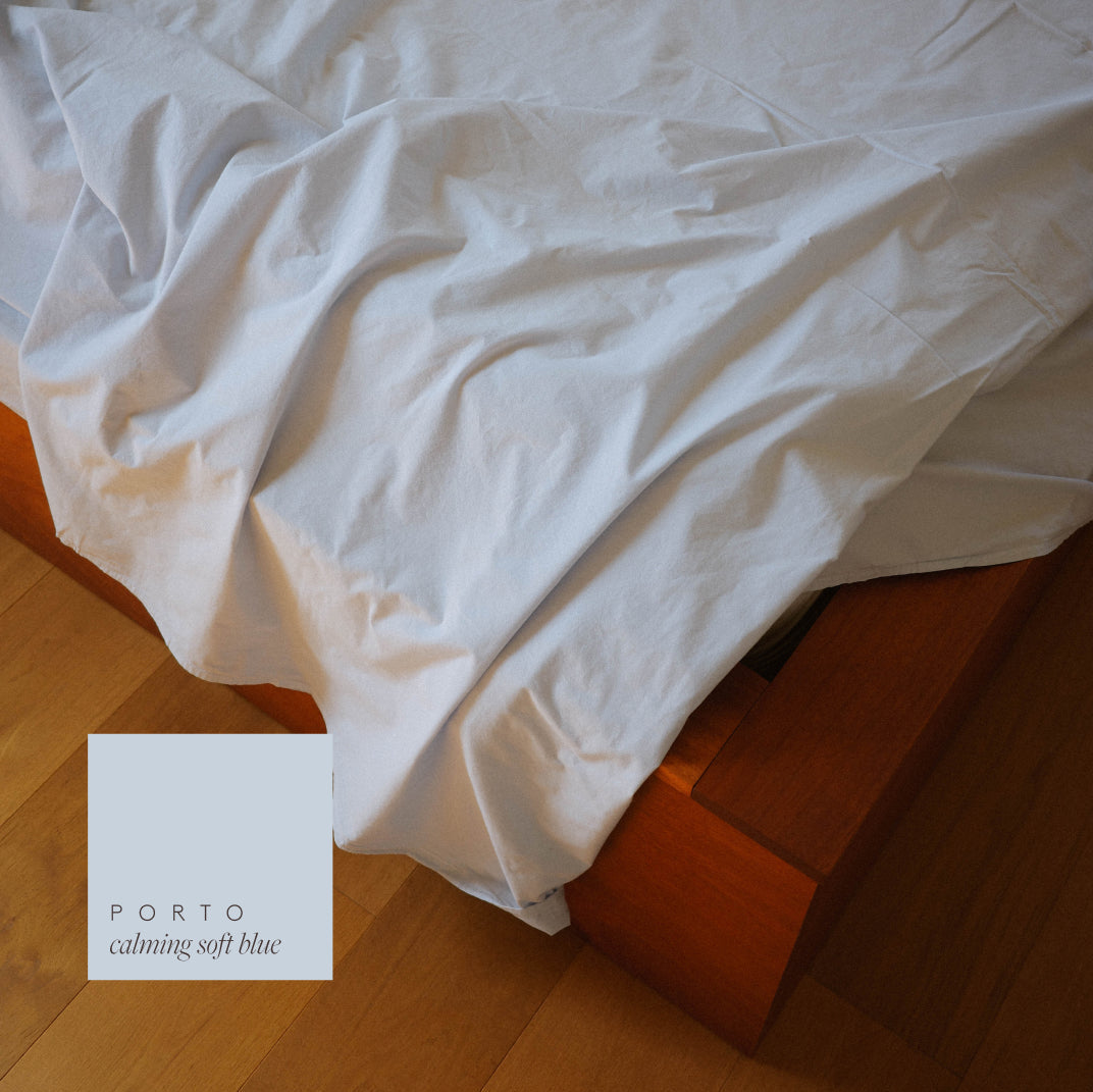 Light blue flat sheet, draped over a wooden bed frame on a light wood floor, with a color swatch label reading 'PORTO calming soft blue' in the bottom-left corner.