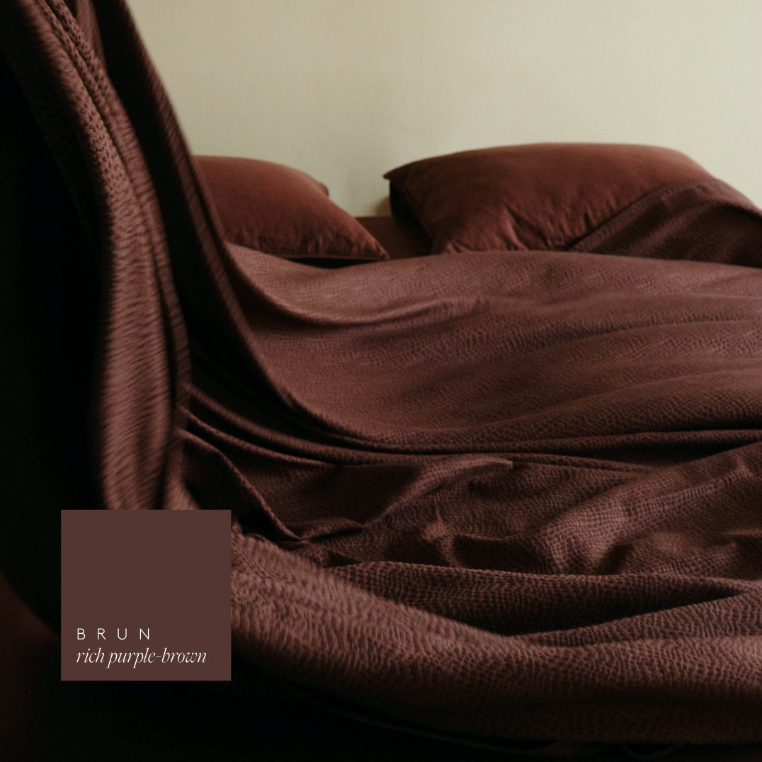 A close-up of a bed made with rich purple-brown bedding, showing a textured duvet cover and matching pillowcases in a warm brown tone. A small label in the lower left reads 'BRUN rich purple-brown'.