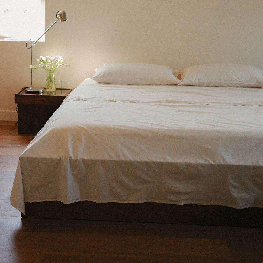 A neatly made bed with white bedding and two white pillows in a softly lit bedroom, with a dark wood nightstand, floor lamp, and vase of flowers beside it.