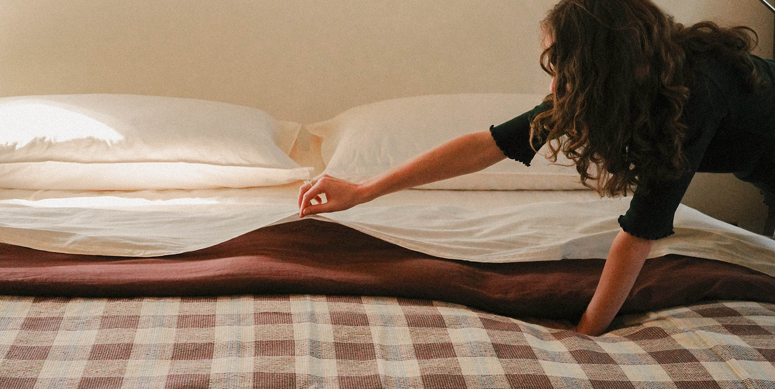Person reaching towards a bed with white sheets and brown plaid blanket.
