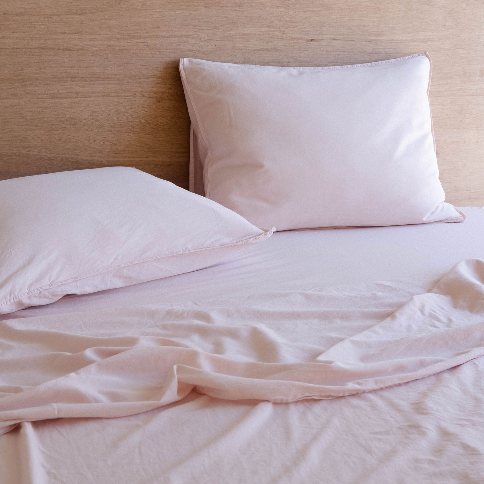 A bed dressed with light pink sheets and pillows against a wood headboard 