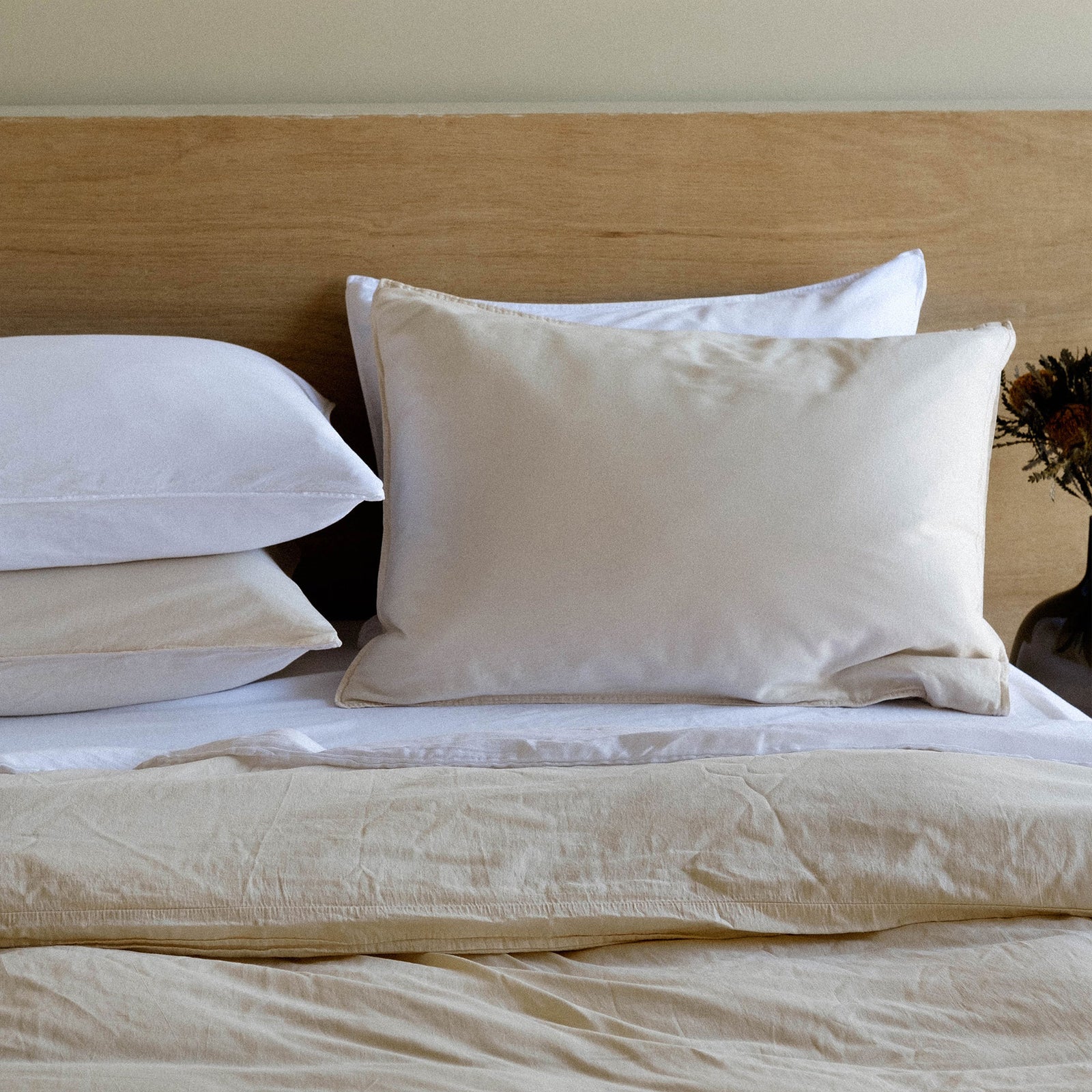 Close up of a bed dressed in cream and beige sheets with stacked pillows against a light wood headboard
