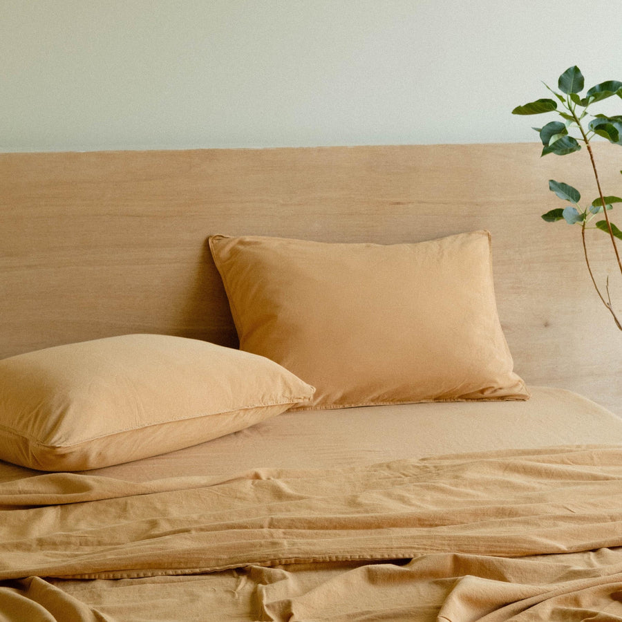 Two light camel matte sateen pillows on a bed with light camel matte sateen sheets against a wooden headboard 