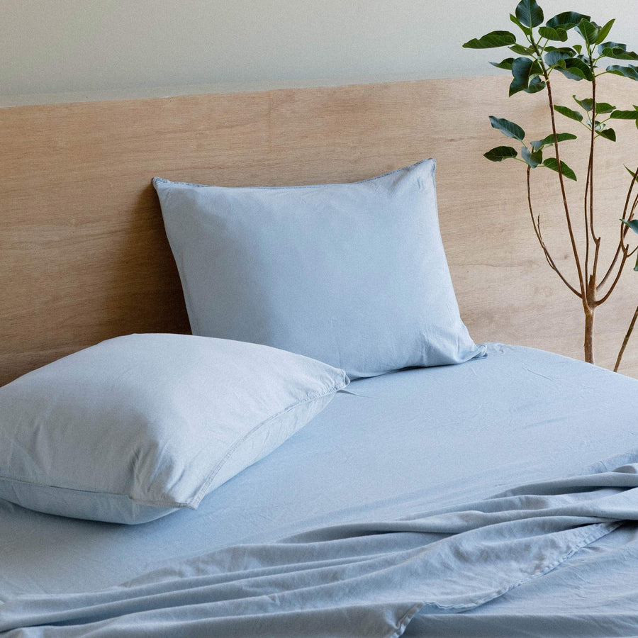 A bed with made with Turkish Blue sheets. Two pillows rest against a wooden headboard and there is a small tree in the background.