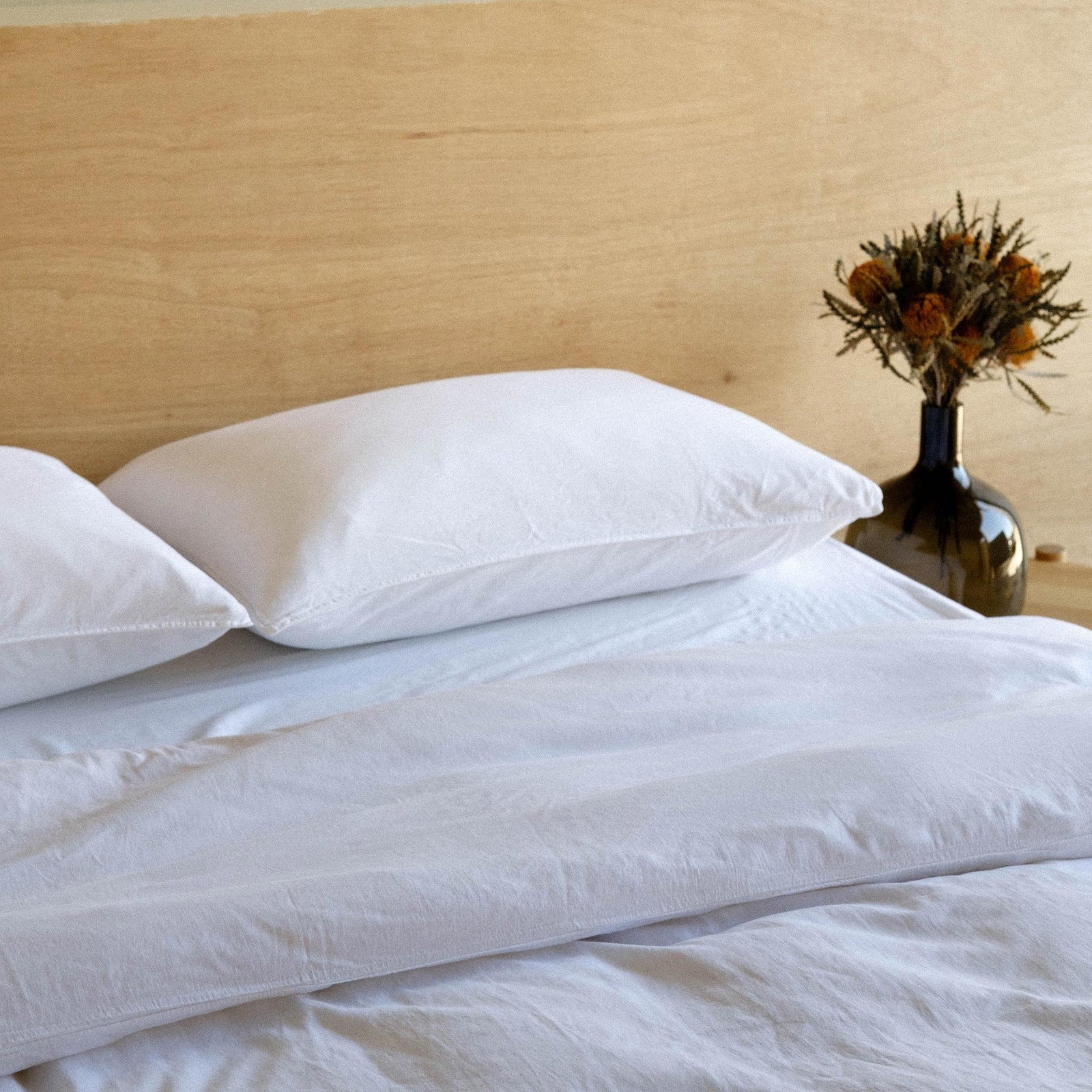 A neatly made bed with white matte sateen sheets and a vase of dried flowers on the nightstand by the bed