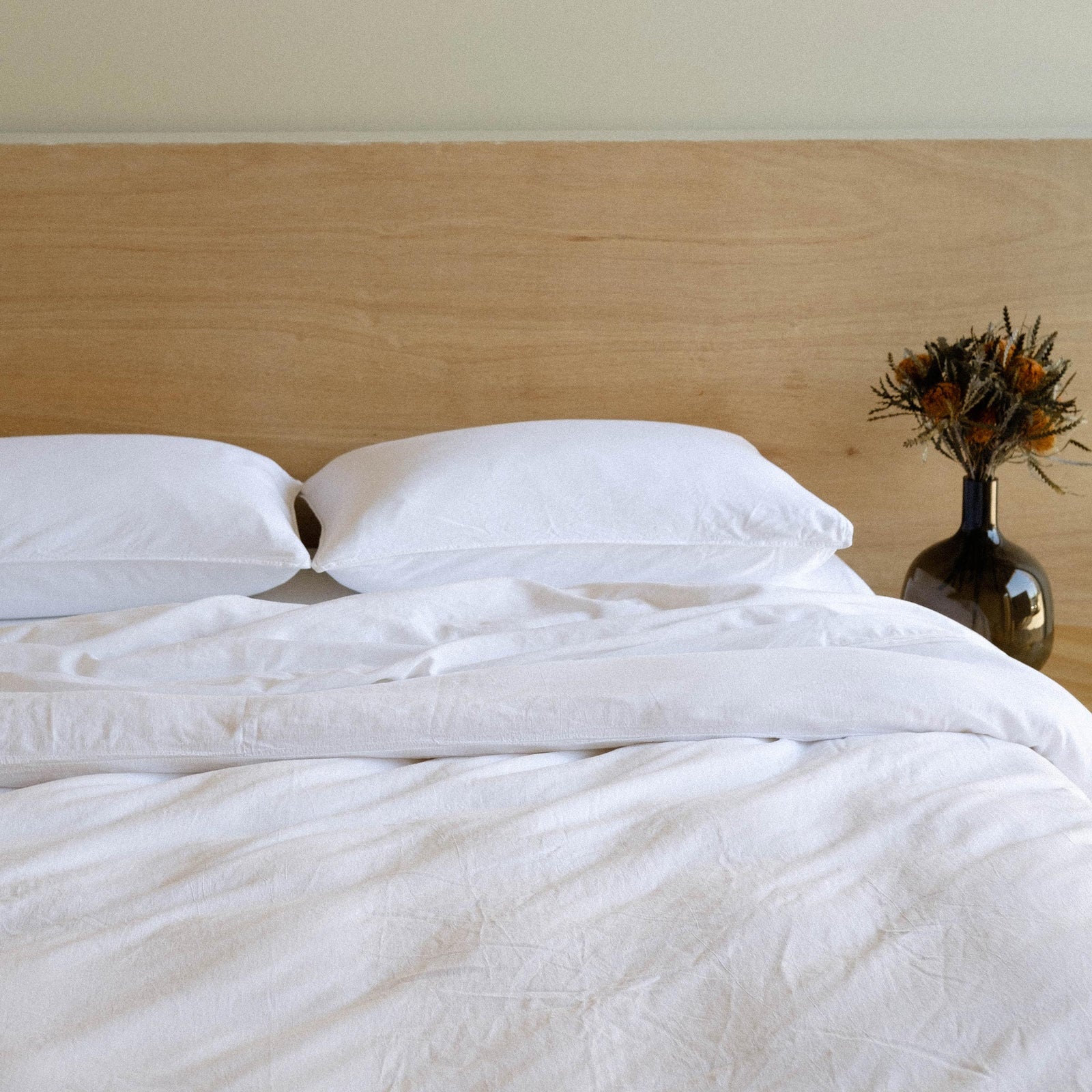 A bed dressed in white matte sateen with two pillows, sheets and a duvet cover in a bedroom setting next to a base of dried flowers